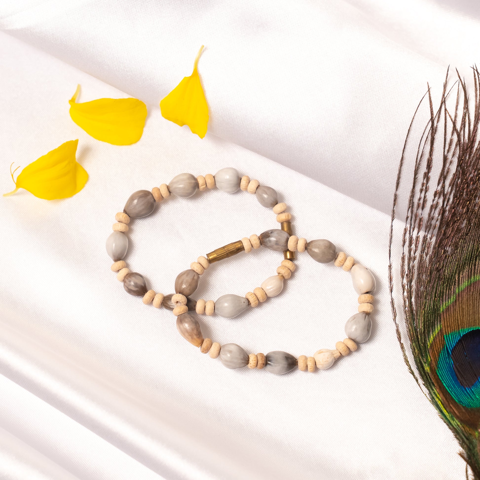 Mala with gray and beige beads on a white surface with yellow leaves and a peacock feather.