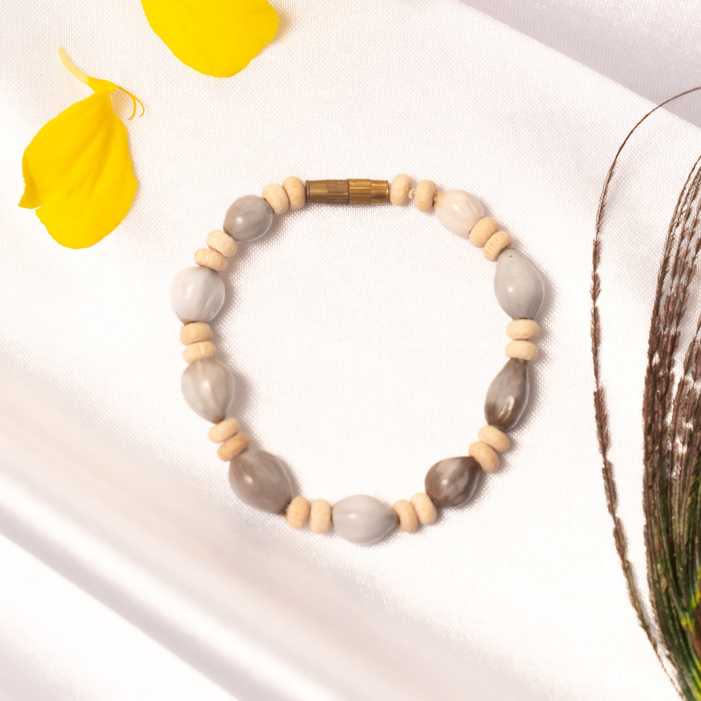 Beaded bracelet with gray and beige beads on a white surface with yellow petals and greenery.